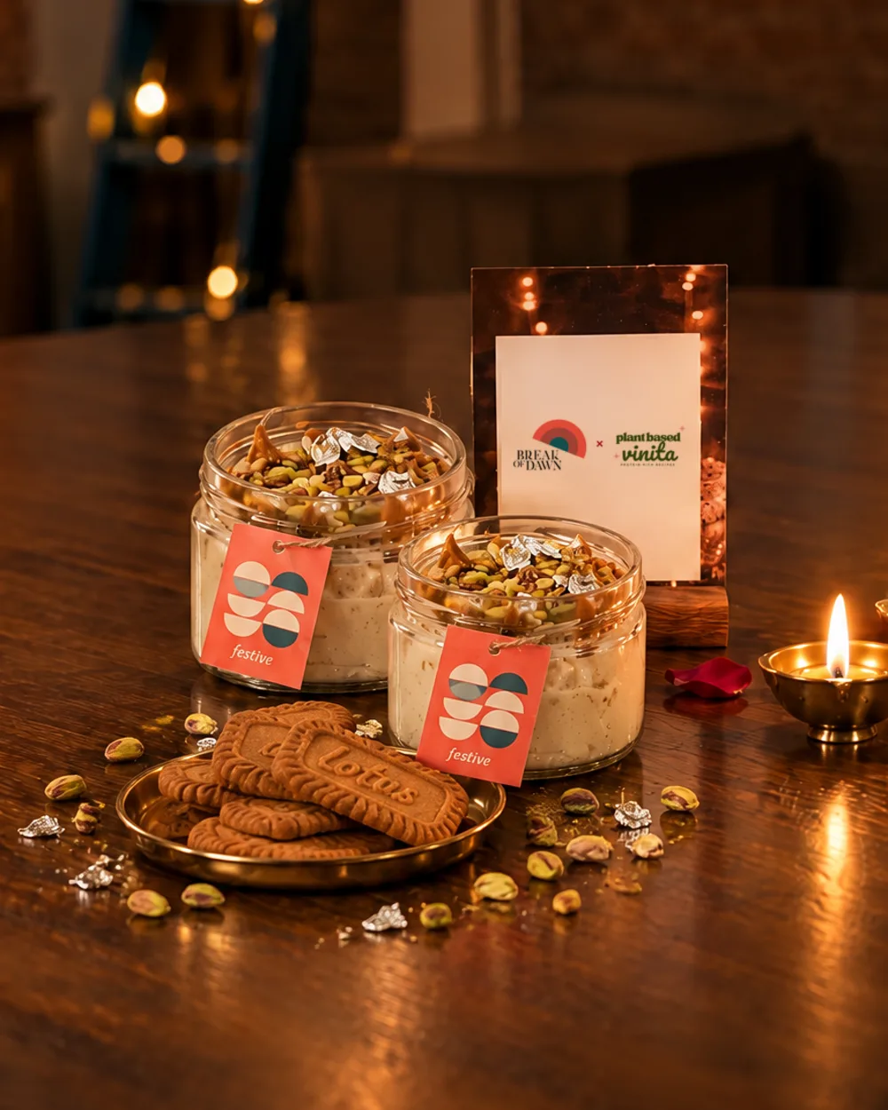 Two jars of Break of Dawn Biscoff kalakand with festive tags, Biscoff biscuits on a brass plate and the Break of Dawn x Plant Based Vinita card