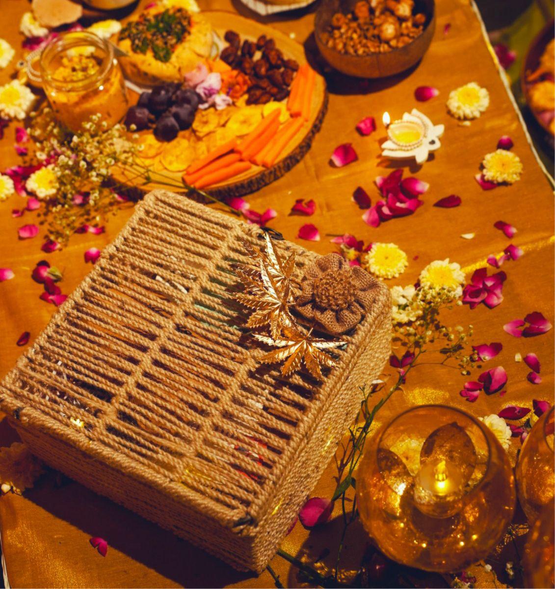 A lidded jute gift box finished with gold leaves and a jute flower, on a gold cloth strewn with rose petals.