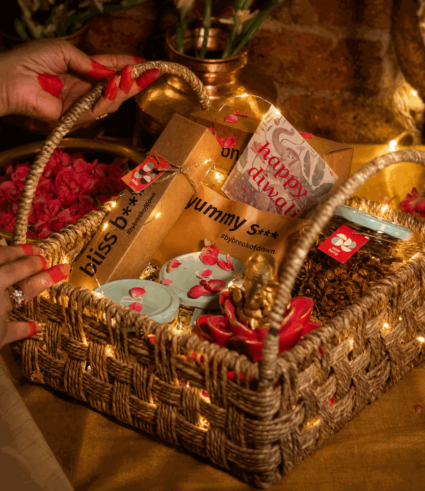 A jute basket being packed with bliss balls, nogurt, a brass diya and a hand-illustrated card, scattered with rose petals.
