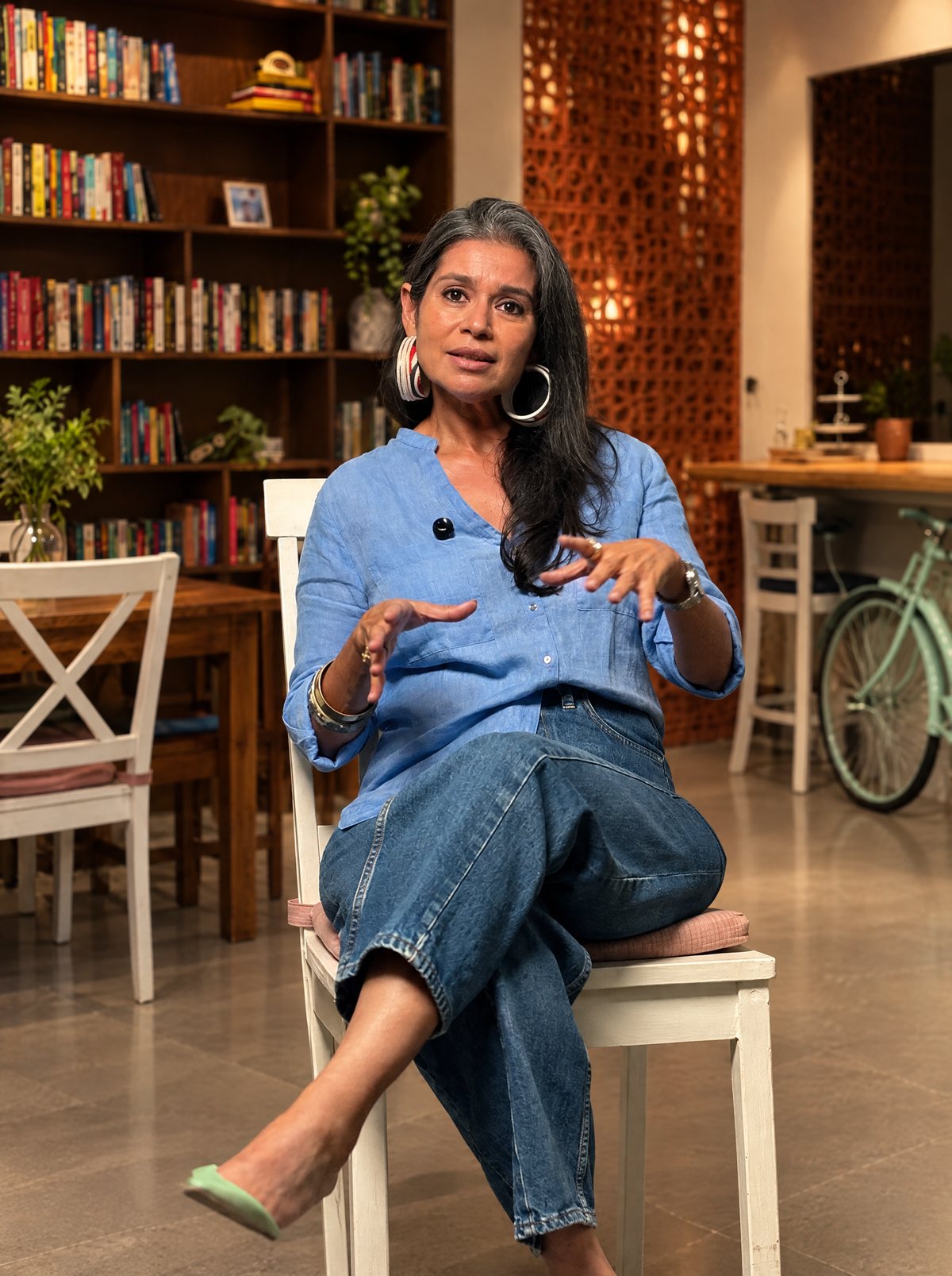 Pardita Mascarenhas, founder of Break of Dawn, mid-sentence on a white chair at Gather, the Break of Dawn studio, with bookshelves and a brick screen behind her